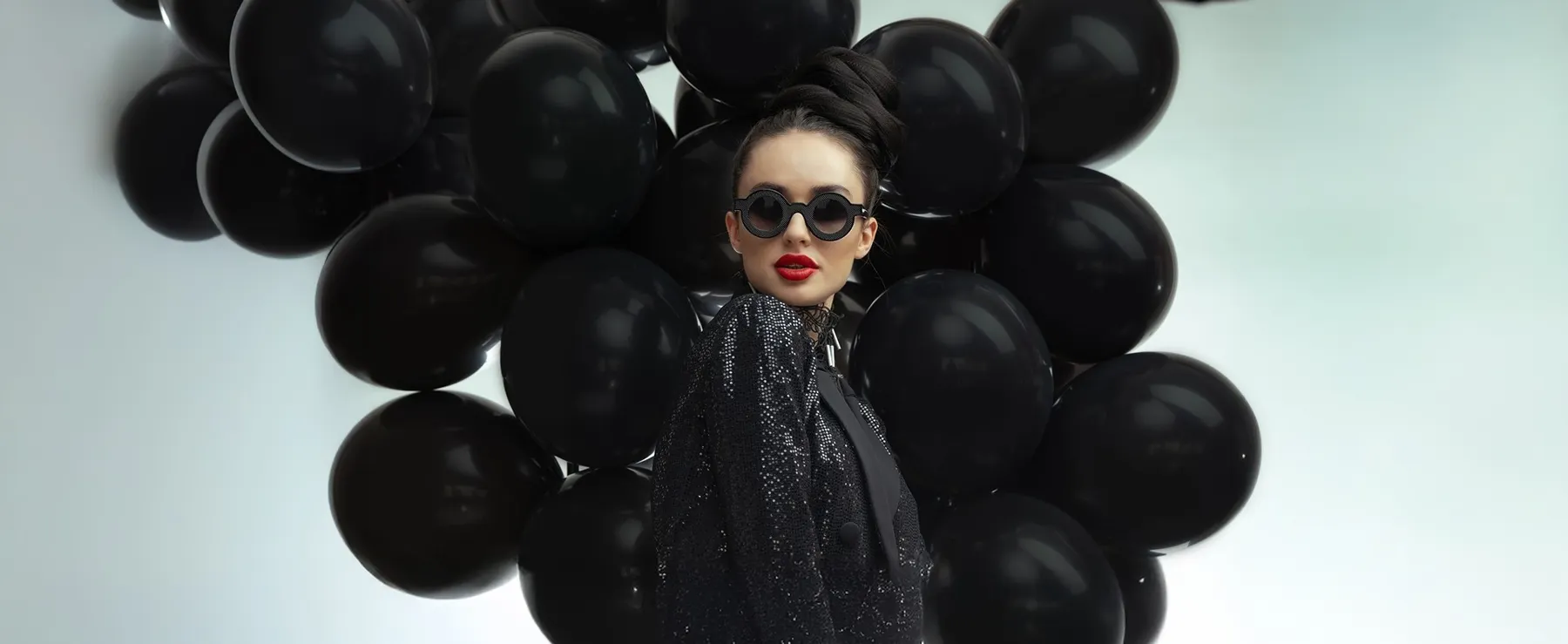 Naočare Sabine Be, fotografija kuće: žena u crnim okruglim sunčanim naočarima, u crnom sakou, među crnim balonima